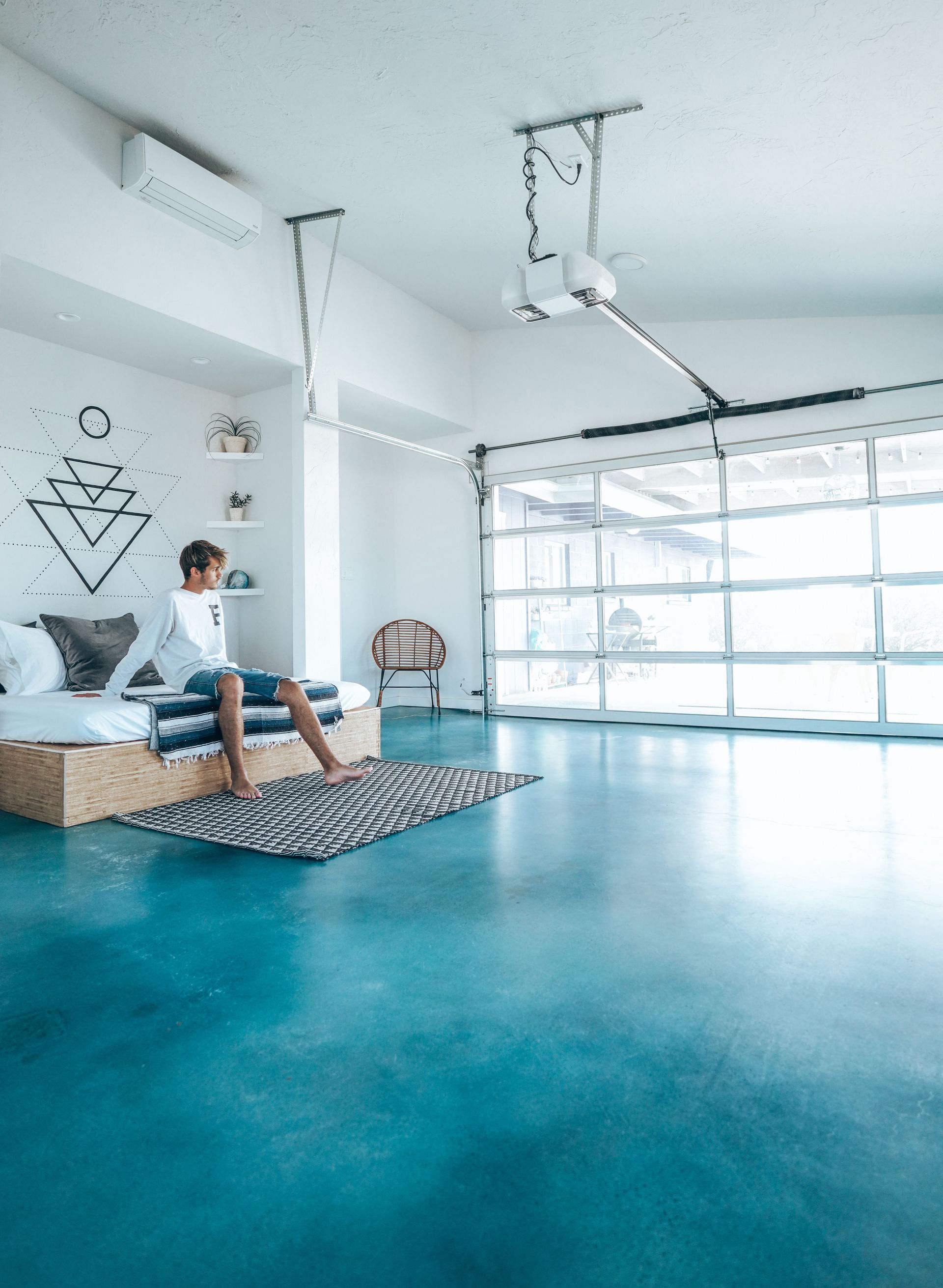 Spacious minimalist bedroom interior with large windows and a man sitting on bed in natural light.